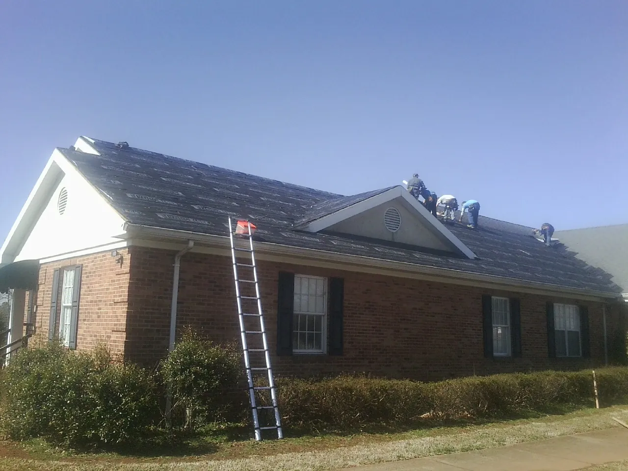 Crew tearing shingles off a brick ranch roof with a ladder set — Metal Roof Repair work by our crew in Dell Rapids